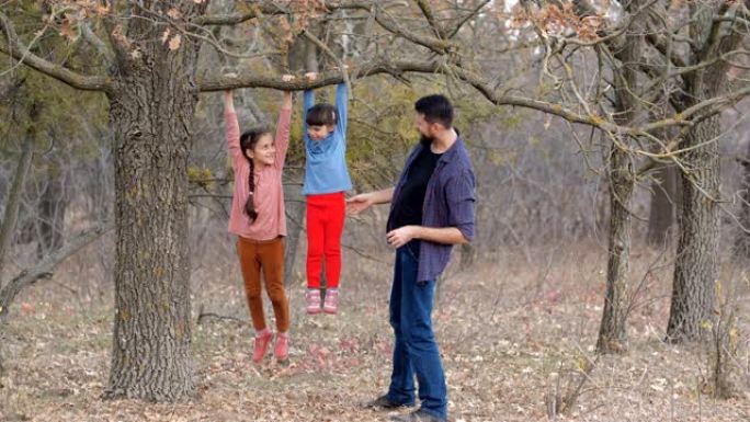 父亲带着两个女儿走在美丽的秋天森林里。穿着格子衬衫的大胡子男人和小女孩玩耍，微笑着，在树上攀爬很开心