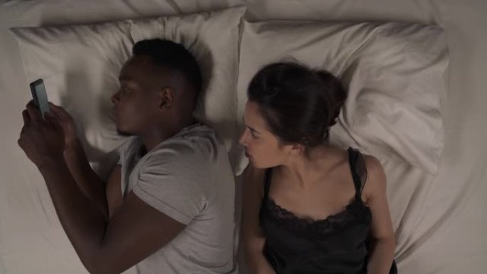 跨种族夫妇晚上躺在床上的俯视图。黑人使用手机。白人女孩从他的肩膀上窥视着屏幕