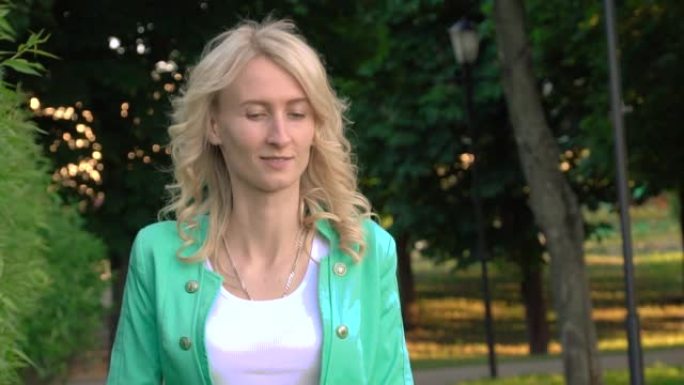 前视图，年轻苗条的高加索金发女郎沿着一条装饰整齐的绿树大道行走。在一个阳光明媚的夏日。慢动作女孩旋转