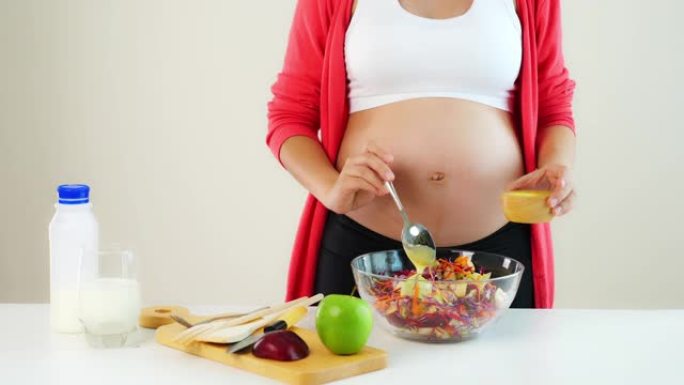 怀孕的年轻女子在厨房的桌子上做沙拉。怀孕的母亲把美味的低脂沙拉酱放在蔬菜沙拉中，用叉子吃沙拉。无法识