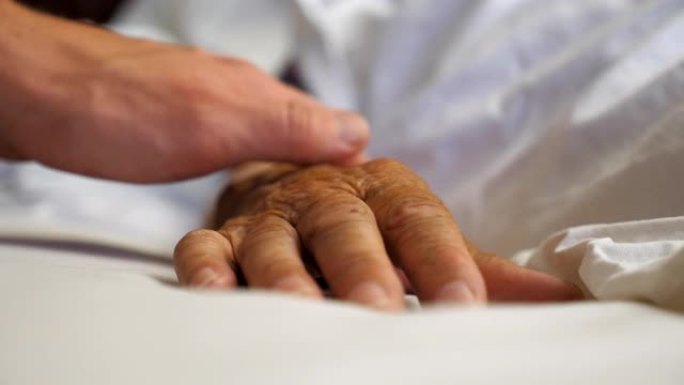 年轻人在诊所轻轻抚摸生病的成熟女人的手。孙子安慰躺在病床上的年迈奶奶皱巴巴的手臂。儿子支持他的老父母