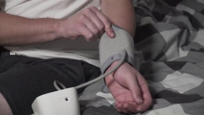 年轻的白人男子在卧室里坐在床上穿着家装测量血压。男性使用医疗器械眼压计检查自己的血压。年轻时的高血压
