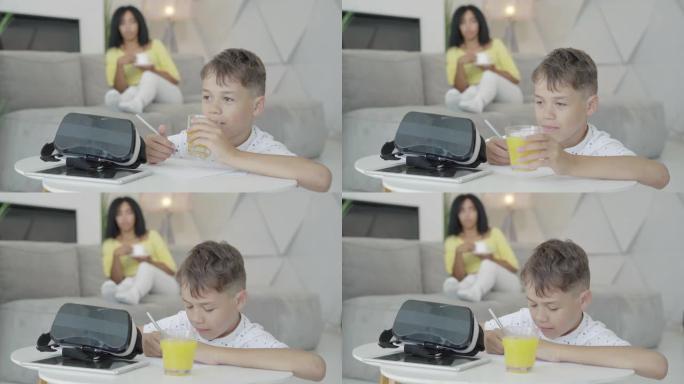 快乐的小男孩喝橙汁和做作业的特写镜头。开朗的非洲裔美国儿子的肖像在家里学习，而模糊的母亲在背景的沙发