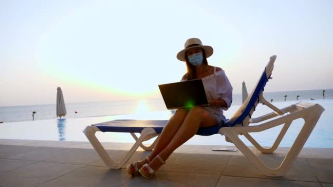 年轻女子戴着防护面具，戴着遮阳帽和夏装，在笔记本电脑上工作，坐在游泳池旁的躺椅上。远程工作。新常态。