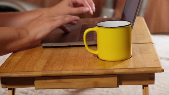 两只女性的手在笔记本电脑键盘上打字，一名技术人员站在一张桌子上，旁边是一个黄色的杯子，上面放着咖啡