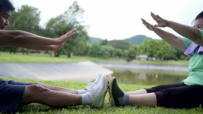 亚洲老年夫妇在公共户外公园锻炼时，手到脚坐在草地上，老年健康退休生活，家庭质量时光，湖山背景