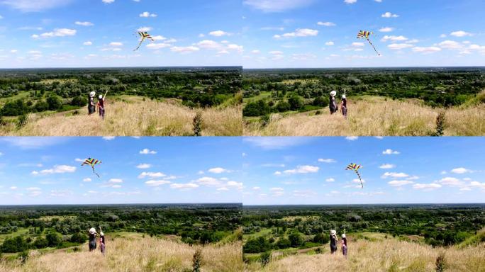 航空。幸福的家庭，妈妈和女儿，在美丽的绿色山谷景观和蓝天的背景下，放飞多色的风筝，玩得开心。后视图。