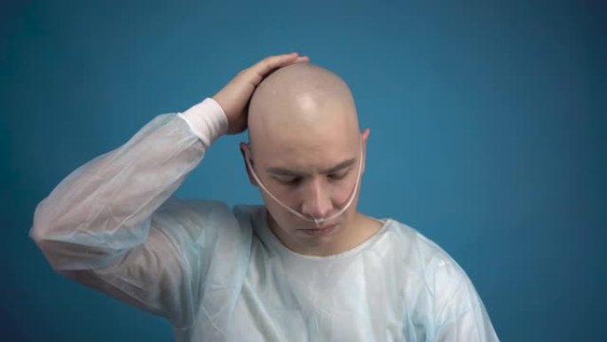 一个秃头的年轻男子带着肿瘤悲伤地看着蓝色背景上的相机。病人摸了摸他的光头。化疗导致脱发。