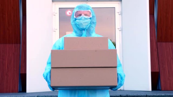 快递员穿着防护服，口罩，护目镜，手里拿着一些纸板箱，在医院门口的背景上。冠状病毒爆发期间的货物运送服