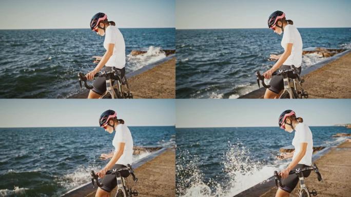 戴着防护头盔，太阳镜和运动服的女孩正坐在海堤的自行车上。她正在用手机打字。晴天，慢动作