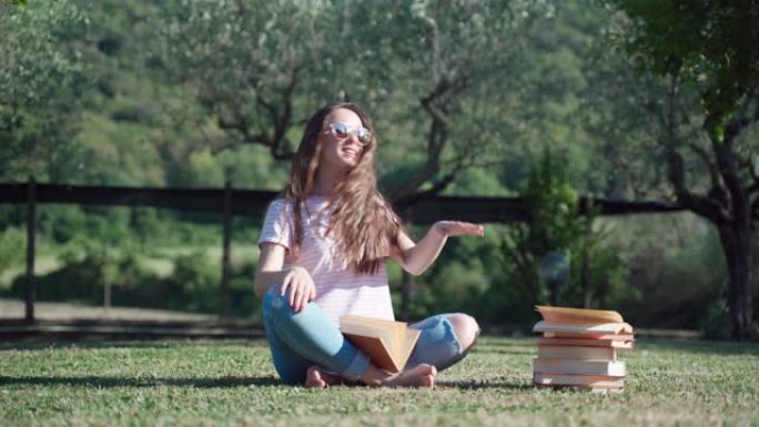 长卷发的微笑女孩坐在大自然全景背景下的草地上跳舞做手部动作。女孩坐在那堆书旁边，从课上休息了。自我教