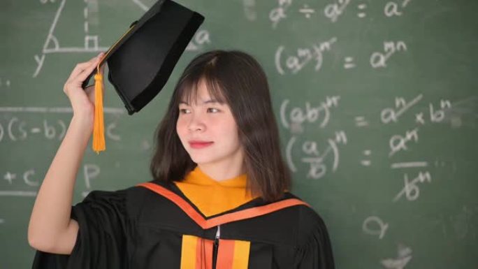 亚洲女毕业生穿着黑色长袍和黑色帽子，并配以金色流苏。站在教室的黑板前