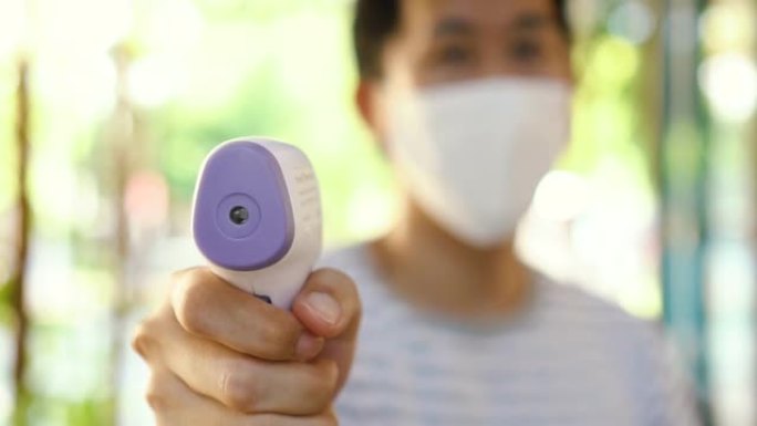 戴着口罩的亚洲年轻人使用医用数字红外温度计检查和监测体温。新的正常生活来筛选访客以防止冠状病毒新型冠