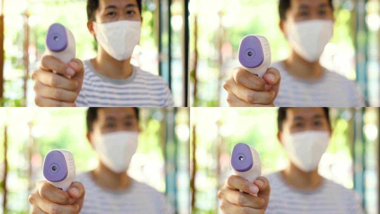 戴着口罩的亚洲年轻人使用医用数字红外温度计检查和监测体温。新的正常生活来筛选访客以防止冠状病毒新型冠
