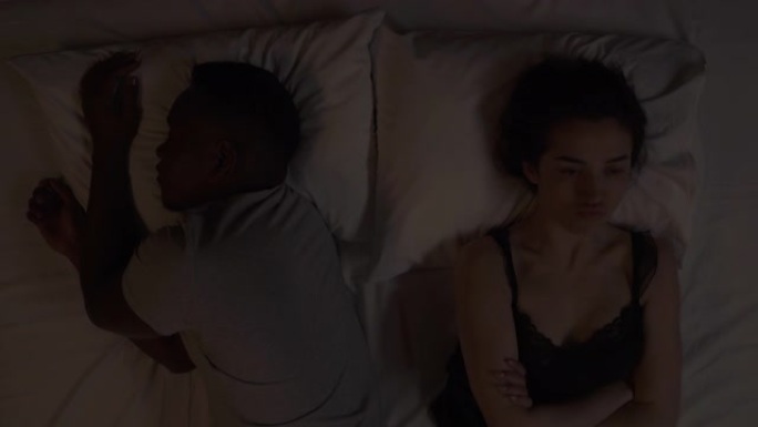 多种族夫妇晚上躺在床上的特写镜头。高加索女孩的景象和摇头，因为她的非洲男友正在睡觉