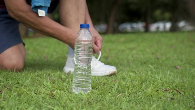 亚洲男性跑步者在绿草地上锻炼时系鞋带，户外公园，积极的生活方式，保持水合水瓶放在一边，积极的人寿保险