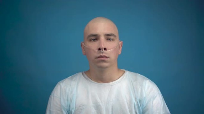 一个秃头的年轻男子带着肿瘤悲伤地看着蓝色背景上的相机。病人拉直呼吸器的管子。化疗导致脱发。