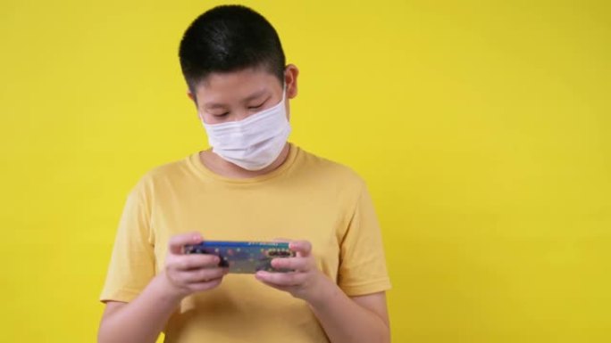 快乐的亚洲青春期男孩戴着口罩，玩黄色背景的网络游戏，新的正常概念。