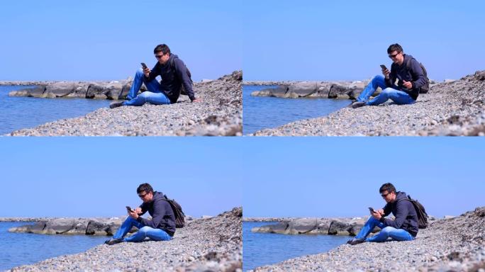 Man traveller坐在海石海滩上，微笑并在智能手机上键入消息。