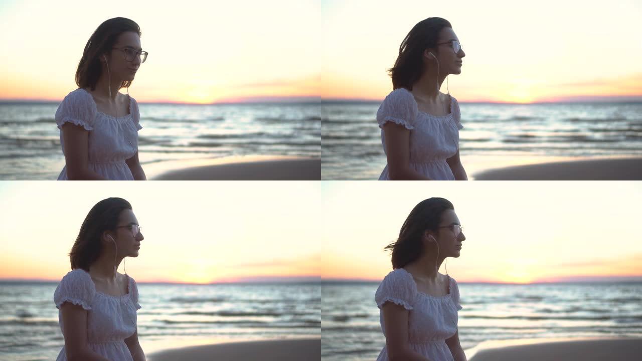 一名年轻女子坐在海边的一块石头上，耳朵里戴着耳机。日落时一个穿着白色连衣裙的女孩在耳机上听音乐特写镜