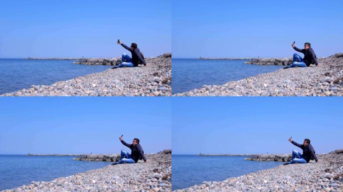Man traveller坐在海石海滩上，在智能手机上自拍。
