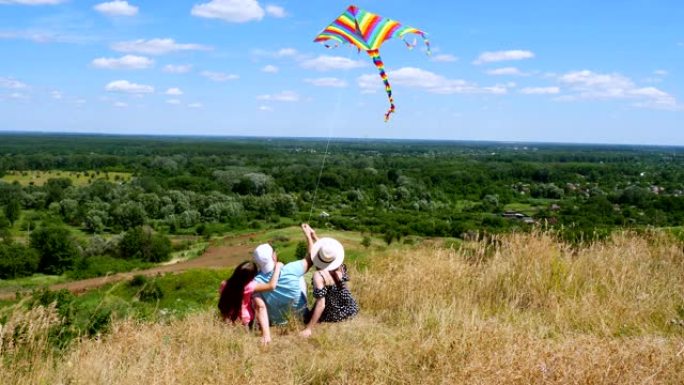 快乐的年轻家庭坐在悬崖边缘，放飞五颜六色的风筝，玩得开心，背景是惊人的绿色山谷全景，蓝天与罕见的云。