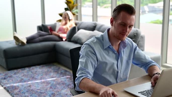 年轻情侣恋人穿着休闲装一起在家里的4K分辨率的客厅笔记本电脑上工作的快乐和放松的生活方式。