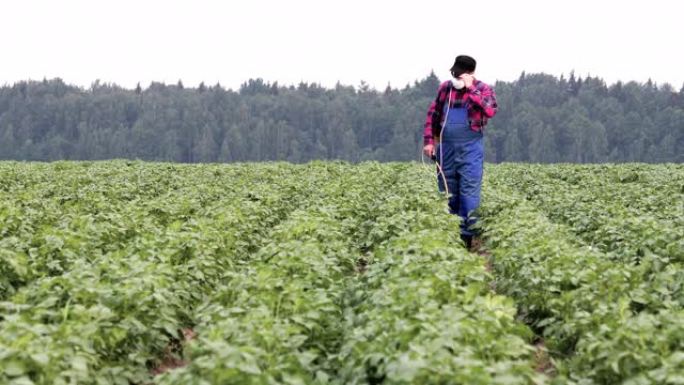 一个年轻的农民在工作服和带有喷雾器的呼吸器在马铃薯田里的肖像