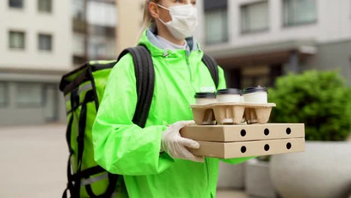 穿着安全口罩，手套和绿色制服的无法识别的女性食品快递员的跟踪镜头，手里拿着披萨盒和外卖咖啡杯，交付订