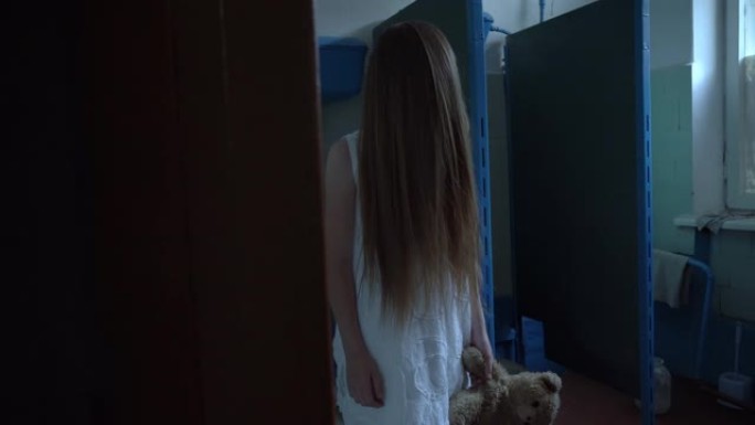 在旧学校里，在厕所里的一个房间里，一个女孩穿着白色连衣裙，脸上留着长发。在他的手中，他有一只泰迪熊，
