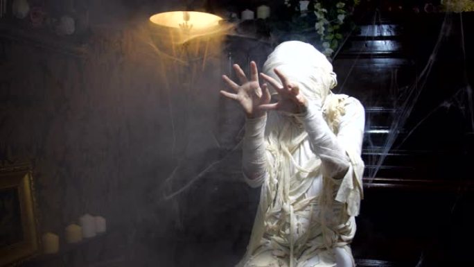 穿着万圣节木乃伊服装的女孩在古老的黑暗哥特式室内摆出姿势并吓scar的电影镜头，上面覆盖着蜘蛛网和雾
