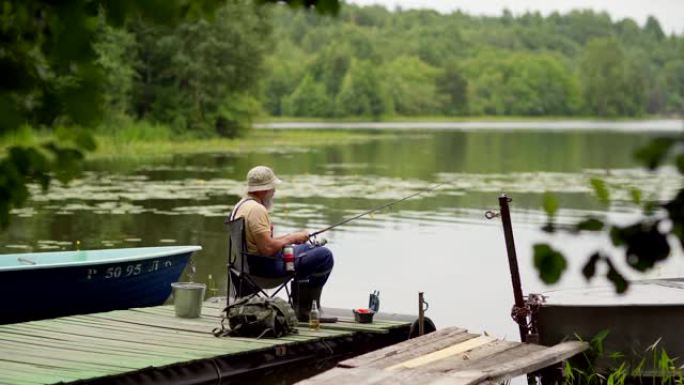 锁定侧视镜头，大胡子的高级男子戴着斗帽钓鱼和喝啤酒，坐在乡村湖边木墩的椅子上