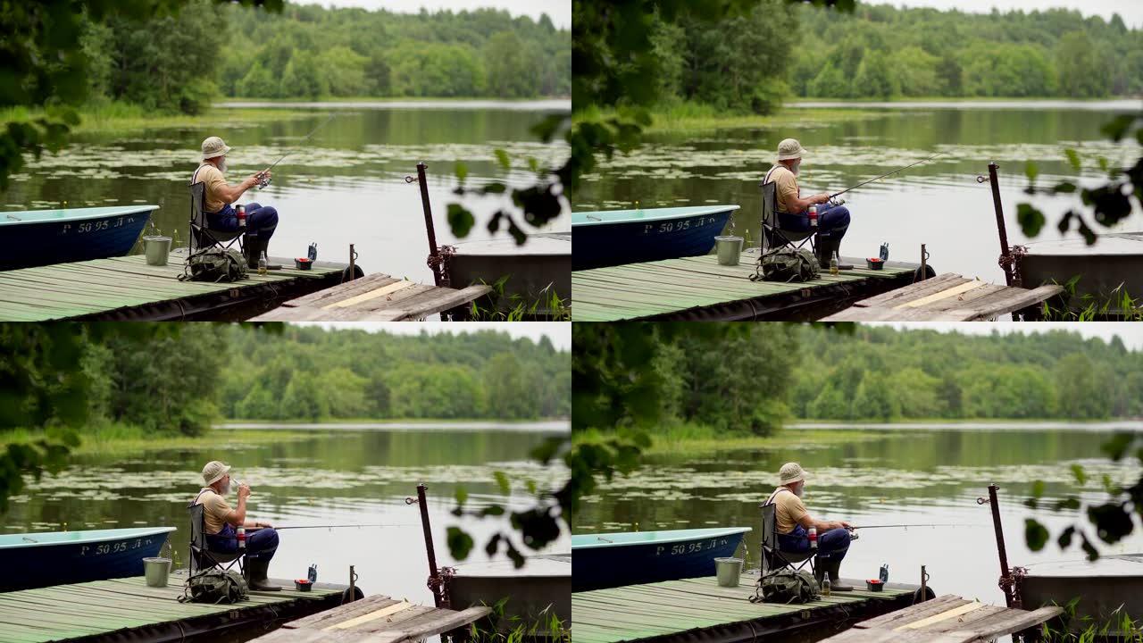 锁定侧视镜头，大胡子的高级男子戴着斗帽钓鱼和喝啤酒，坐在乡村湖边木墩的椅子上