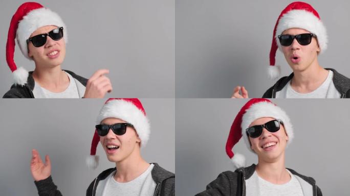 圣诞少年男孩问候歌曲叮当太阳镜圣诞帽唱快乐家庭看起来相机灰色背景。圣诞气氛和歌曲，传统。寒假。复制空