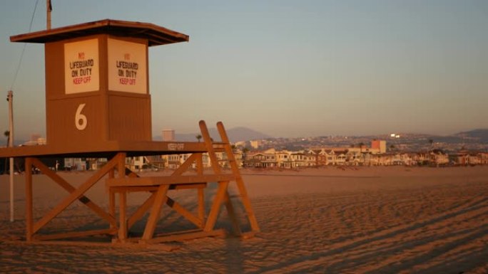 标志性的复古木制橙色救生员守望塔在沙质加利福尼亚太平洋海滩上被日落光线照亮。私人度假屋和地平线上的山