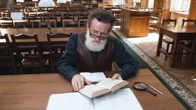 有吸引力的人专注于阅读大胡子的老人眼镜坐在图书馆的桌子旁看书