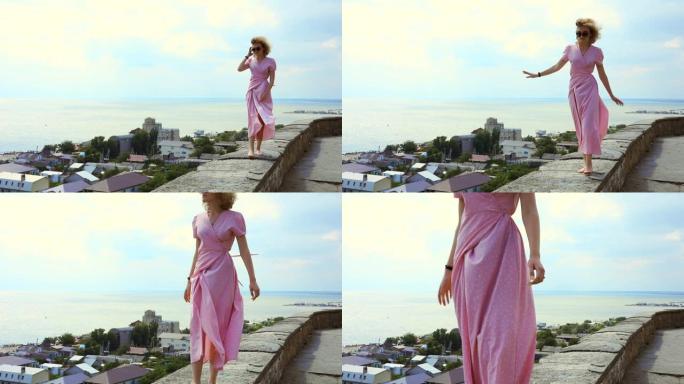 穿着粉红色连衣裙的女人在大海的背景下沿着露台散步