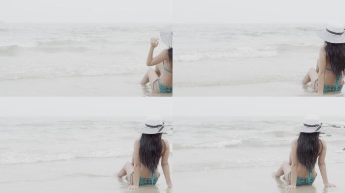 穿着比基尼和帽子的美丽的年轻亚洲女性在度假时坐在沙滩上享受乐趣，女孩休闲，在暑假里用水浪在海上放松，