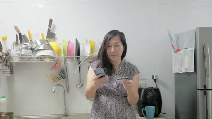 亚洲妇女使用信用卡在家里的厨房在线购物，生活方式的概念。