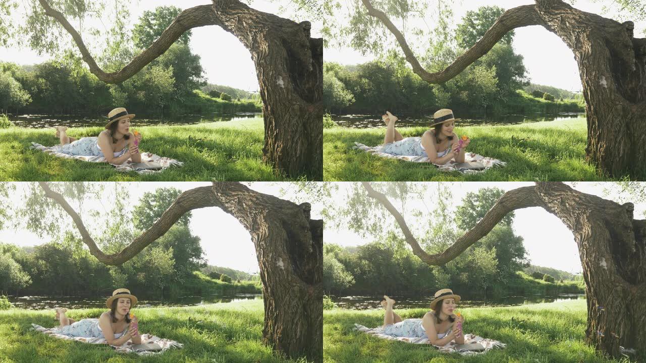 年轻可爱的女孩在大树下的草地上的毯子上放松，在阳光明媚的夏日吹泡泡。穿着帽子和衣服的迷人女性在河边的