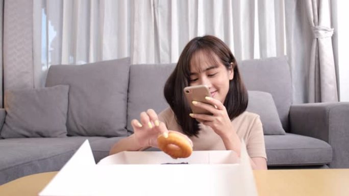 年轻的亚洲女性外卖吃甜甜圈垃圾食品享受自拍在家里的客厅社交更新，快餐送货服务安全，阻止冠状病毒通过社