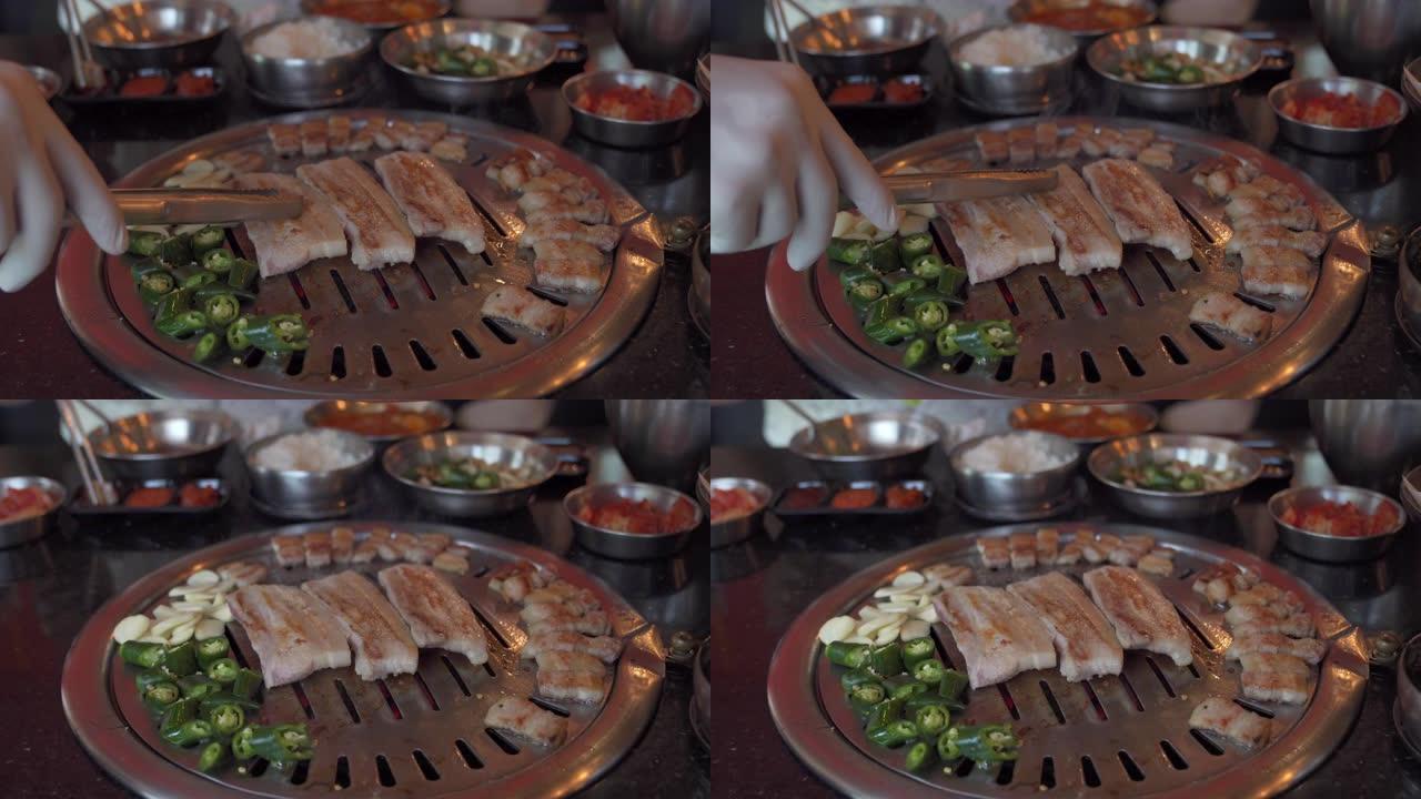 在餐厅的韩国铁烧烤烤盘上，厨师手工烹饪熏烤猪肚肉和大蒜和青辣椒的4k特写镜头。传统韩国风格的食物烧烤