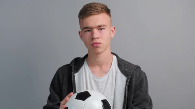 十几岁的男孩的肖像站在他的手中，在他面前严肃地拿着足球，然后在电视上观看足球比赛。足球迷，球员。青少