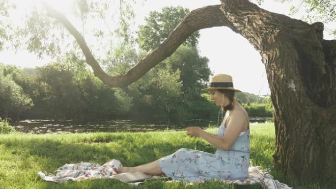 迷人的年轻女子坐在河边公园的格子上，戴上耳机。阳光明媚的夏日，女孩一边享受耳机音乐，一边在大树下的公