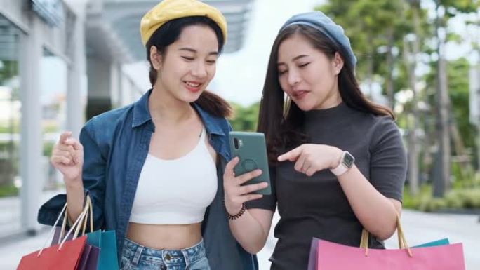 微笑的年轻亚洲女人，购物中心背景上的彩色包。使用智能手机在线购物，站在商场大楼时微笑。生活方式概念。
