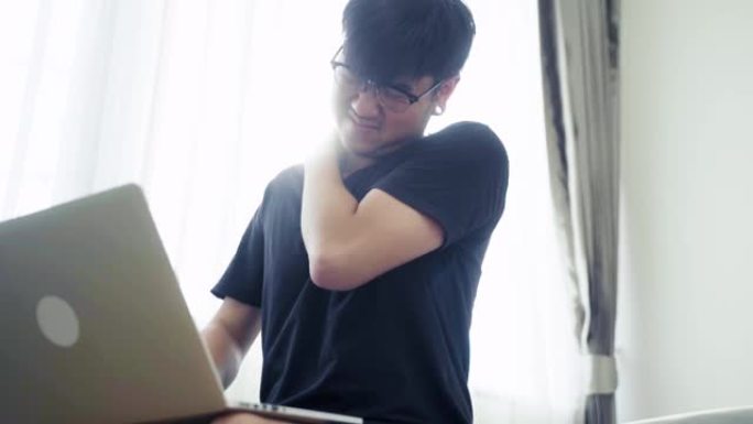 一个戴着玻璃的男人在计算机上工作，他的肩膀疼痛，并用手触摸他的应该，转头试图减轻疼痛。慢动作镜头。