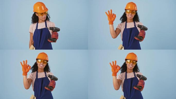 戴着安全帽、护目镜和手套的黑人女性工人。拿着钻头，露出好的手势，微笑着。在蓝色背景上摆姿势