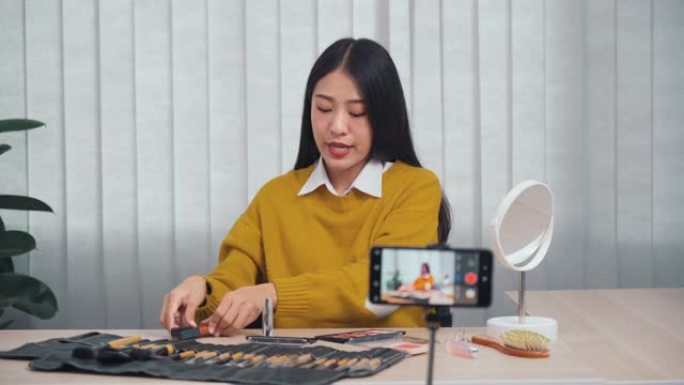 年轻的亚洲女性在家中使用手机摄像头为自己的化妆品博客制作视频，以了解美容产品。