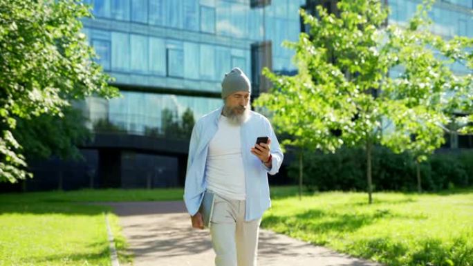 跟踪照片的时尚高级商人，留着灰色胡须，手里拿着笔记本电脑走在绿色城市街道上，在阳光明媚的夏日用手机接