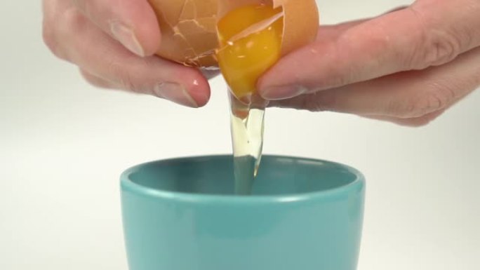 明亮的黄色蛋黄从厨师手中的破碎蛋壳流入蓝色陶瓷杯。
